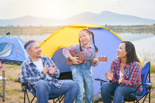 Happy little asian girl playing ukulele and her parents clapping hands at camping site.