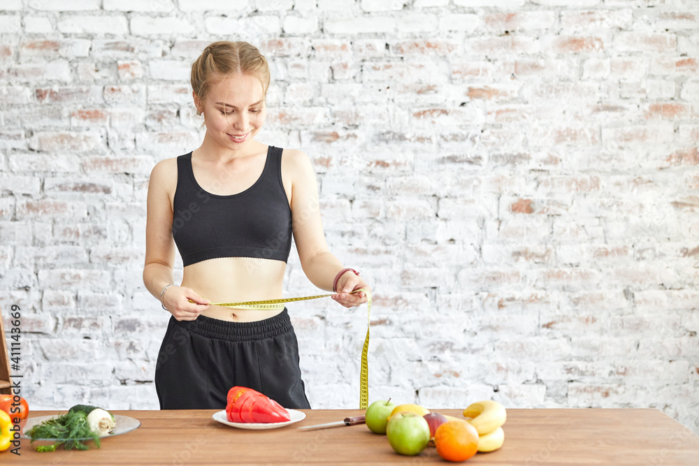 Young woman on a diet, she measures her waist with a soft measuring tape, fresh vegetables on the table Young woman on a diet, she measures her waist with a soft measuring tape, fresh vegetables on the table