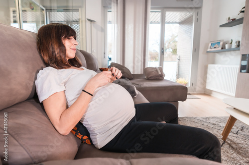 Young pregnant woman with gestational diabetes self-test to control sugar on the sofa at home, measuring the drop of blood from her finger