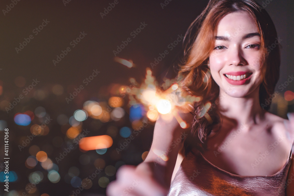 Beautiful young asian brunette woman with white dress joying Christmas or New Year night on a city.Woman holding sparkler against defocused lights, close up.