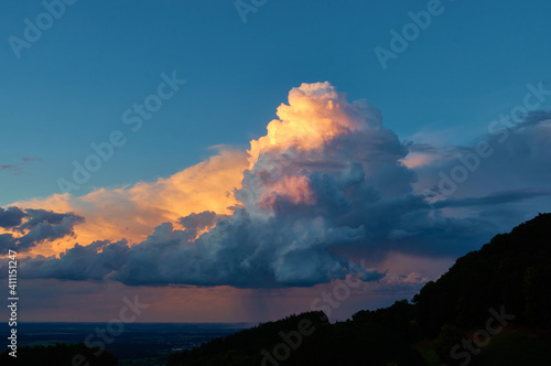 Large white weather cloud glows orange in the evening light, blue sky. trees as silouhette. Germany