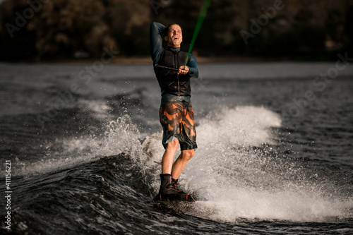 Fototapeta Naklejka Na Ścianę i Meble -  young man in black life jacket holds bright green rope and rides wakeboard