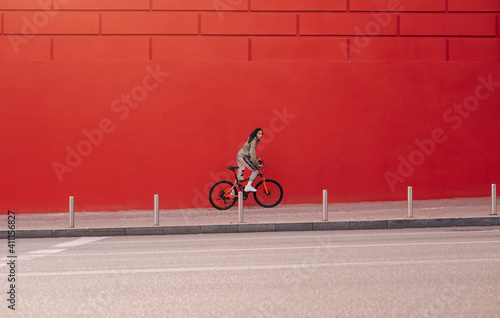 Sporty woman in casual clothes rides a bicycle on a city street on a background of a red wall and looks at the camera. Weekend cycling as a sport