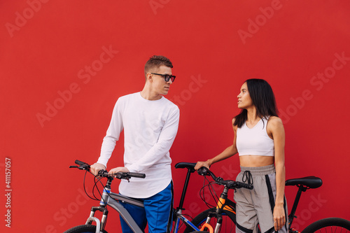 Beautiful couple man and woman in casual clothes with bicycles isolated on red wall background, resting after riding and smiling.