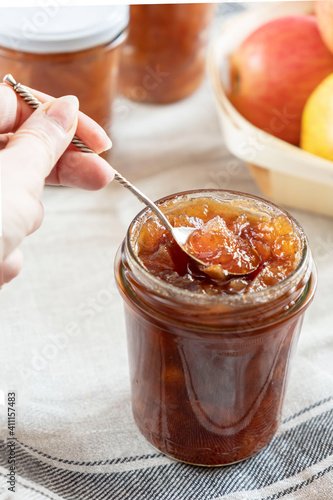 Homemade apple jam marmalade with cloves and cinnamon from organic apples in a transparent glass jar with vintage retro spoon in woman female hand. Homemade autumn jam preserves.