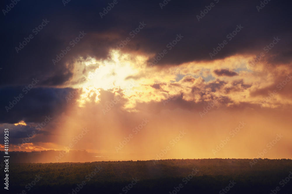 Sunbeams make their way through black rain clouds over trees and silhouettes of city buildings Sunbeams make their way through black rain clouds over trees and silhouettes of city buildings