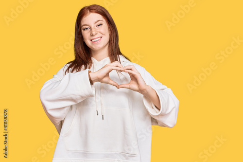 Young read head woman wearing casual sweatshirt smiling in love showing heart symbol and shape with hands. romantic concept.