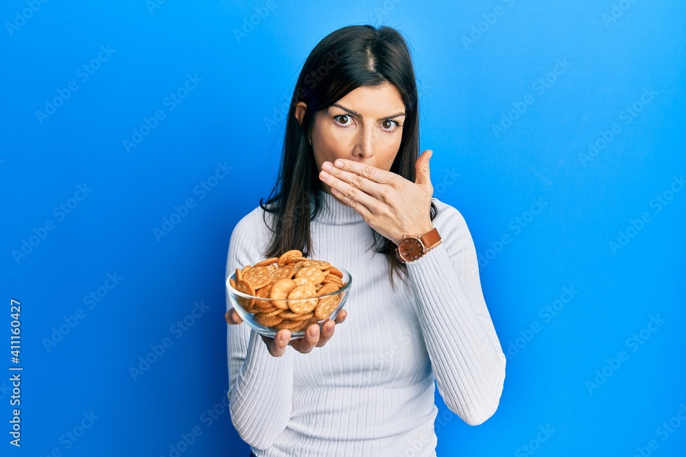Young hispanic woman holding salty biscuits bowl covering mouth with hand, shocked and afraid for mistake. surprised expression Young hispanic woman holding salty biscuits bowl covering mouth with hand, shocked and afraid for mistake. surprised expression