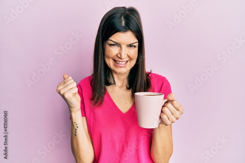 Young hispanic woman drinking a cup of coffee screaming proud, celebrating victory and success very excited with raised arms