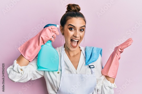 Beautiful brunette young woman wearing apron holding cleaning spray pointing to head celebrating victory with happy smile and winner expression with raised hands