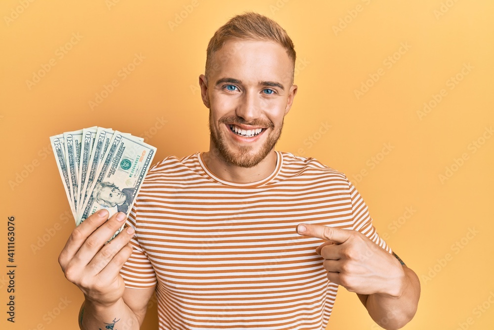 Young caucasian man holding dollars smiling happy pointing with hand and finger Young caucasian man holding dollars smiling happy pointing with hand and finger