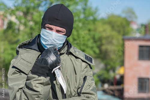 Pensive criminal with a knife in a medical mask and balaclava squints his eyes because of the bright sun © Николай Чекалин