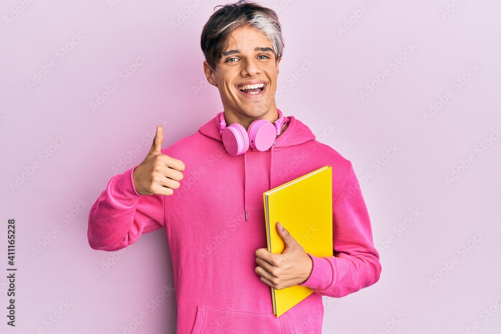 Young hispanic man holding book using headphones smiling happy and positive, thumb up doing excellent and approval sign Young hispanic man holding book using headphones smiling happy and positive, thumb up doing excellent and approval sign