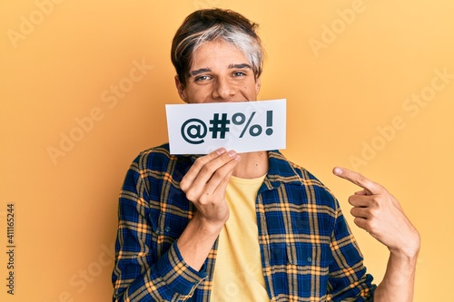 Young hispanic man covering mouth with insult message paper smiling happy pointing with hand and finger