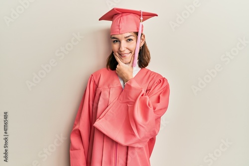 Young caucasian woman wearing graduation cap and ceremony robe looking confident at the camera smiling with crossed arms and hand raised on chin. thinking positive.