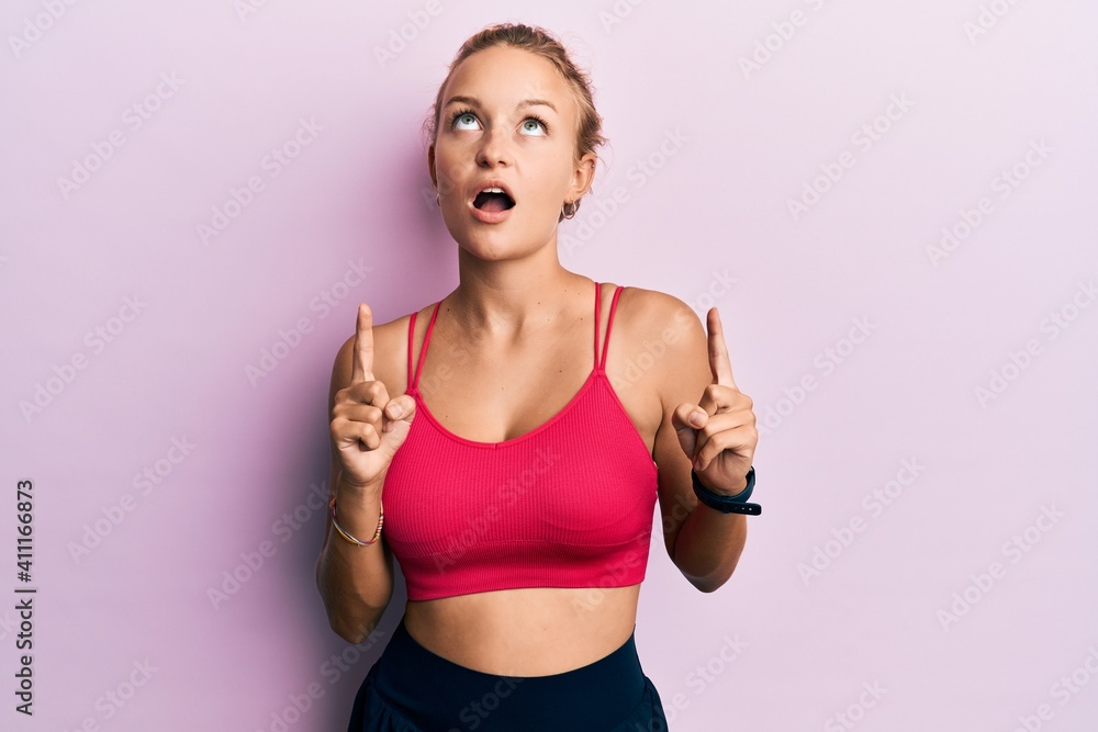 Beautiful caucasian woman wearing sportswear amazed and surprised looking up and pointing with fingers and raised arms. Beautiful caucasian woman wearing sportswear amazed and surprised looking up and pointing with fingers and raised arms.