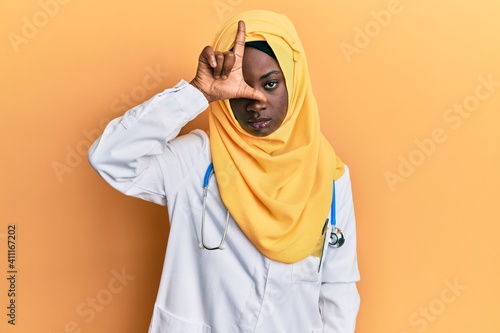 Beautiful african young woman wearing doctor uniform and hijab making fun of people with fingers on forehead doing loser gesture mocking and insulting.
