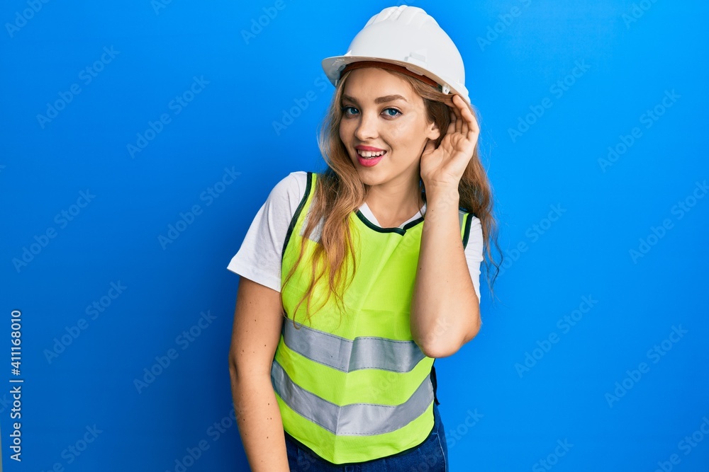 Beautiful blonde caucasian woman wearing architect hardhat smiling with hand over ear listening and hearing to rumor or gossip. deafness concept. Beautiful blonde caucasian woman wearing architect hardhat smiling with hand over ear listening and hearing to rumor or gossip. deafness concept.