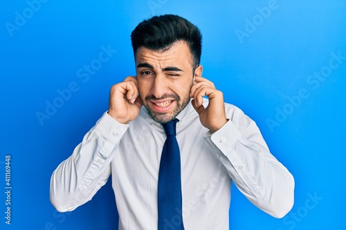 Young hispanic man wearing business clothes covering ears with fingers with annoyed expression for the noise of loud music. deaf concept.