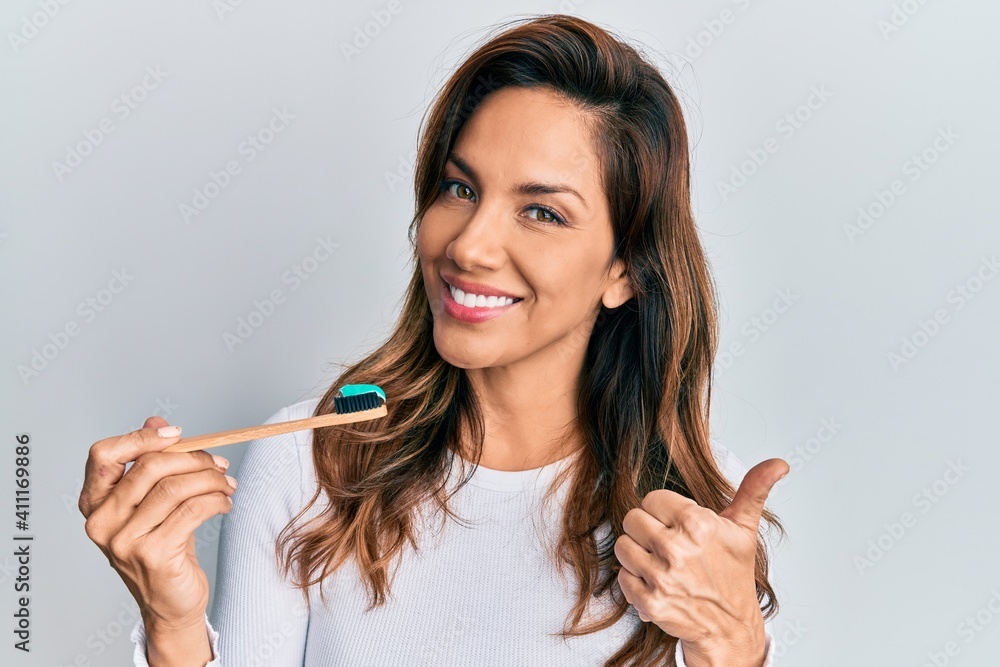 Young latin woman holding toothbrush with toothpaste smiling happy and positive, thumb up doing excellent and approval sign Young latin woman holding toothbrush with toothpaste smiling happy and positive, thumb up doing excellent and approval sign