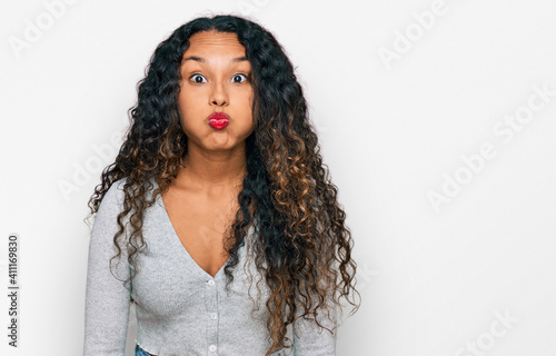 Young hispanic woman with curly hair wearing casual clothes puffing cheeks with funny face. mouth inflated with air, crazy expression.