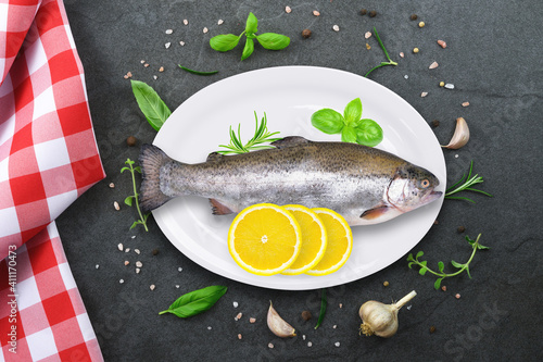 Top view of raw rainbow trout on white ceramic plate on vintage black stone kitchen countertop with  red checkered tablecloth and selection of herbs and spices.