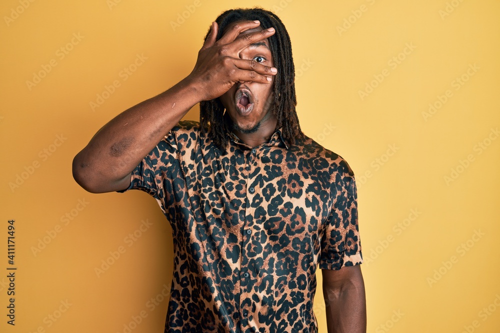 African american man with braids wearing leopard animal print shirt peeking in shock covering face and eyes with hand, looking through fingers with embarrassed expression. African american man with braids wearing leopard animal print shirt peeking in shock covering face and eyes with hand, looking through fingers with embarrassed expression.