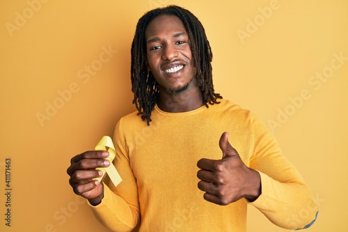 African american man with braids holding suicide prevention yellow ribbon smiling happy and positive, thumb up doing excellent and approval sign photo