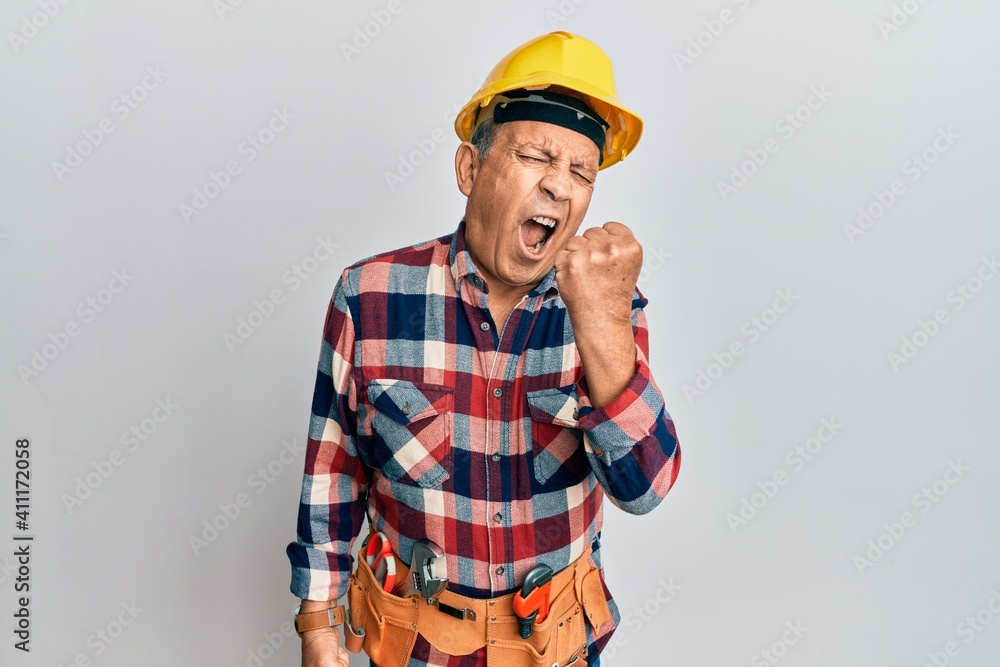 Senior hispanic man wearing handyman uniform angry and mad raising fist frustrated and furious while shouting with anger. rage and aggressive concept.