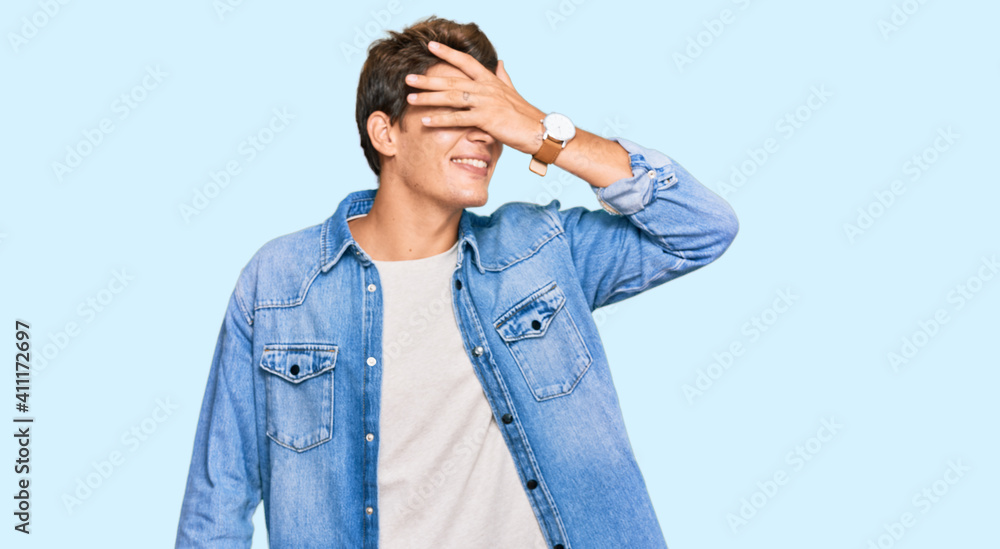 Handsome caucasian man wearing casual denim jacket smiling and laughing with hand on face covering eyes for surprise. blind concept. Handsome caucasian man wearing casual denim jacket smiling and laughing with hand on face covering eyes for surprise. blind concept.