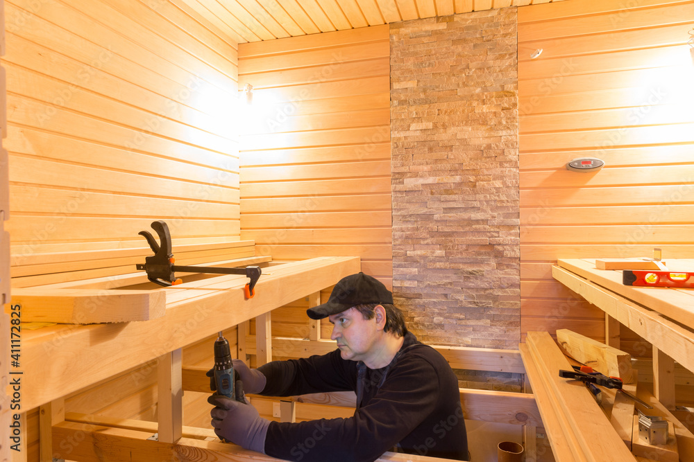 Sauna construction, finishing. The man is screwing a wooden bench to the wall. - obrazy, fototapety, plakaty 