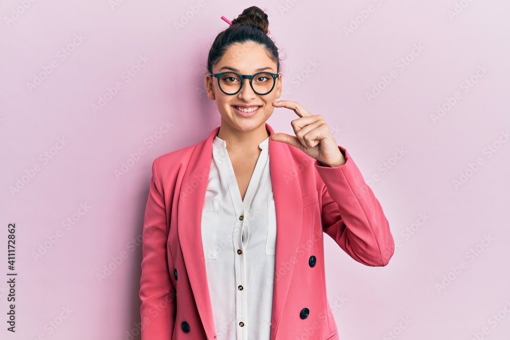 Beautiful middle eastern woman wearing business jacket and glasses smiling and confident gesturing with hand doing small size sign with fingers looking and the camera. measure concept.