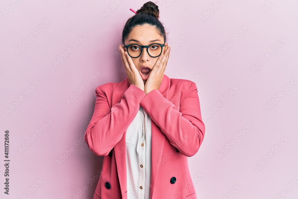Beautiful middle eastern woman wearing business jacket and glasses afraid and shocked, surprise and amazed expression with hands on face
