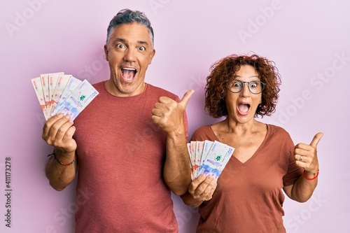 Beautiful middle age couple holding colombian pesos banknotes pointing thumb up to the side smiling happy with open mouth