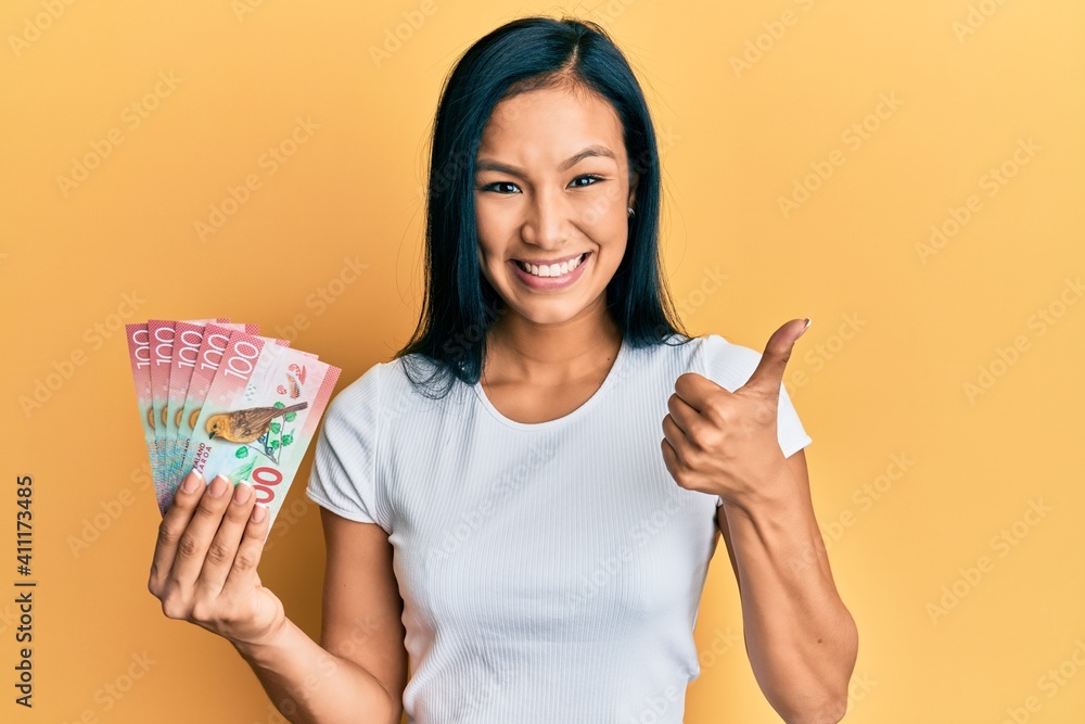 Beautiful hispanic woman holding 100 new zealand dollars banknote smiling happy and positive, thumb up doing excellent and approval sign Beautiful hispanic woman holding 100 new zealand dollars banknote smiling happy and positive, thumb up doing excellent and approval sign