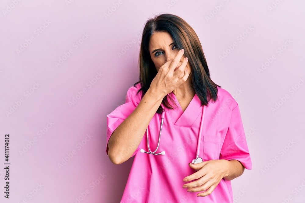 Middle age brunette woman wearing doctor uniform and stethoscope smelling something stinky and disgusting, intolerable smell, holding breath with fingers on nose. bad smell Middle age brunette woman wearing doctor uniform and stethoscope smelling something stinky and disgusting, intolerable smell, holding breath with fingers on nose. bad smell