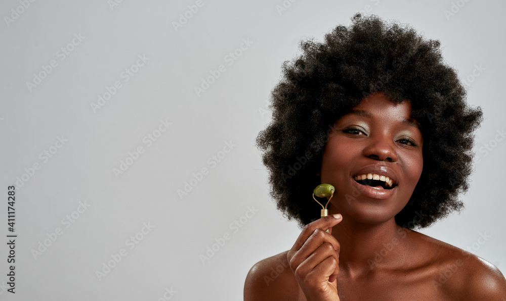 Portrait of excited young african american woman with perfect glowing skin smiling at camera while massaging her face with jade roller, posing isolated over gray background - obrazy, fototapety, plakaty 