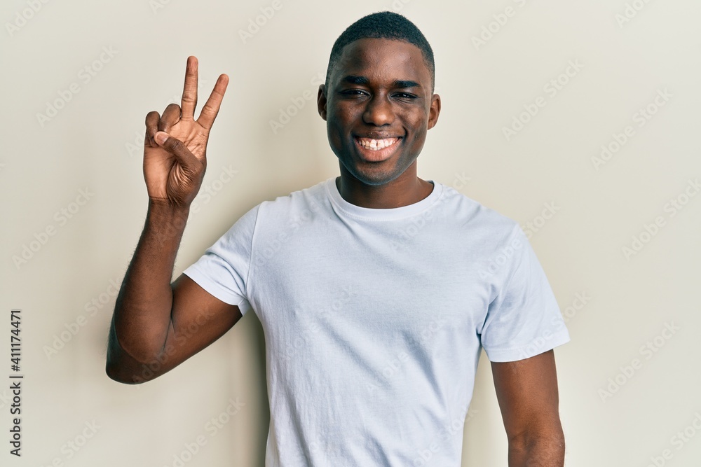 Young african american man wearing casual white t shirt smiling with happy face winking at the camera doing victory sign. number two. Young african american man wearing casual white t shirt smiling with happy face winking at the camera doing victory sign. number two.