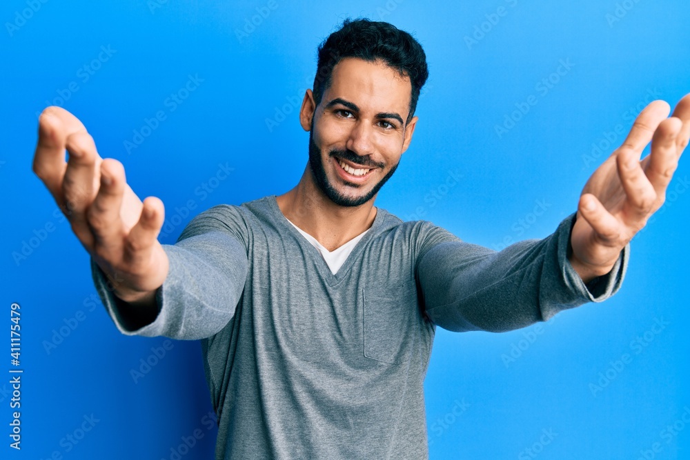 Young hispanic man wearing casual white t shirt looking at the camera smiling with open arms for hug. cheerful expression embracing happiness. Young hispanic man wearing casual white t shirt looking at the camera smiling with open arms for hug. cheerful expression embracing happiness.