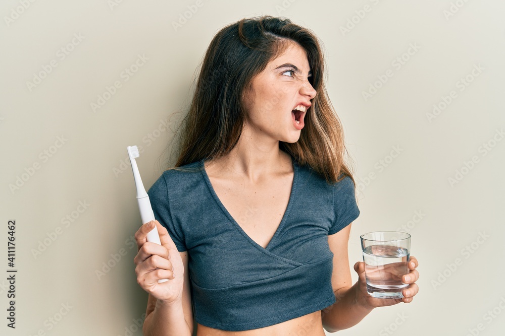 Young caucasian woman holding electric toothbrush and water glass angry and mad screaming frustrated and furious, shouting with anger. rage and aggressive concept. Young caucasian woman holding electric toothbrush and water glass angry and mad screaming frustrated and furious, shouting with anger. rage and aggressive concept.