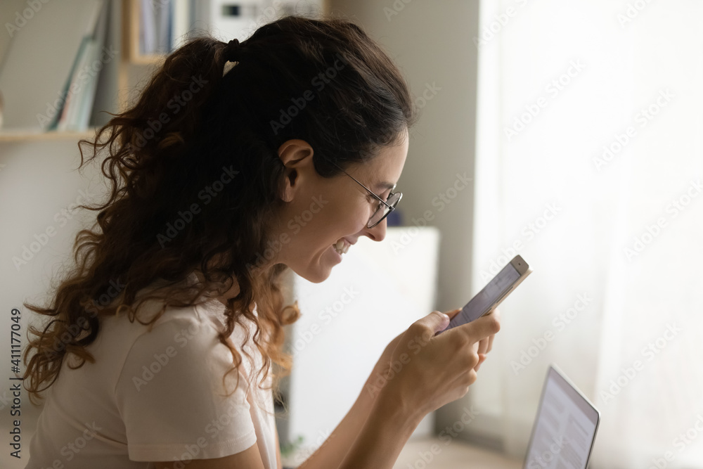 Side view of happy smiling young lady holding mobile phone reading good news receiving visa grant reward. Pleasant woman relax from work at home office chatting messaging online using cell having fun Side view of happy smiling young lady holding mobile phone reading good news receiving visa grant reward. Pleasant woman relax from work at home office chatting messaging online using cell having fun