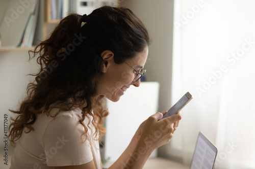 Side view of happy smiling young lady holding mobile phone reading good news receiving visa grant reward. Pleasant woman relax from work at home office chatting messaging online using cell having fun