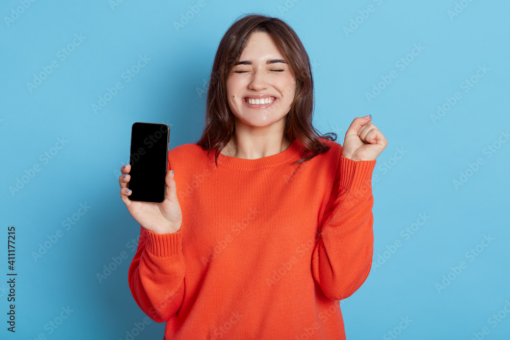 Overjoyed attractive woman keeps fist clenched, being happy to get good news, gets notification on cellphone, wears orange sweater, stands with closed eyes indoor against blue wall, feels amused. Overjoyed attractive woman keeps fist clenched, being happy to get good news, gets notification on cellphone, wears orange sweater, stands with closed eyes indoor against blue wall, feels amused.