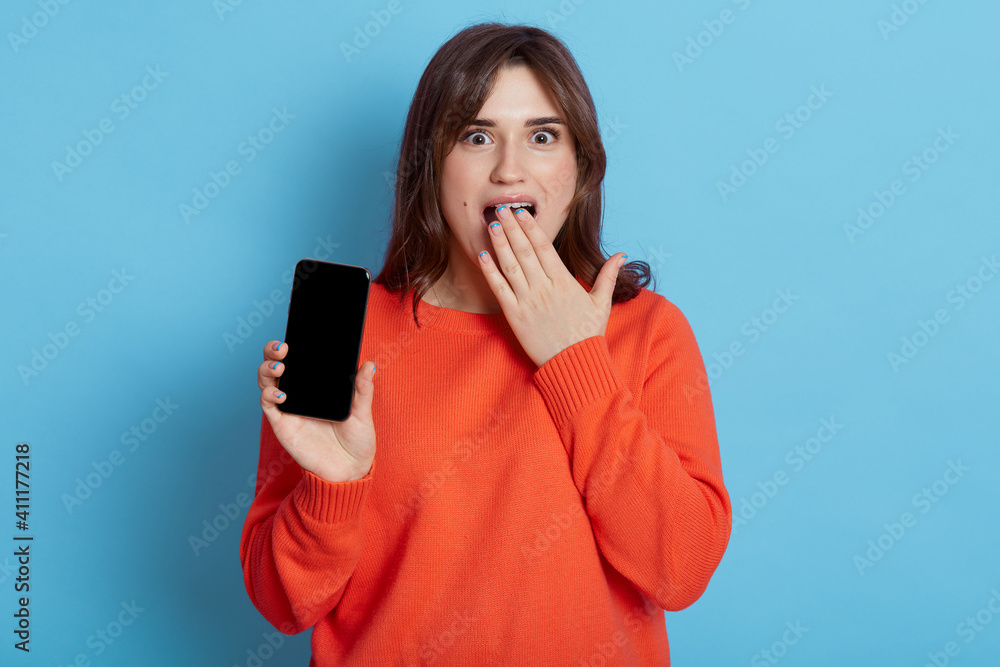 Surprised dark haired woman wears orange sweater, holds mobile phone, gets message with shocking news, looks with amazed expression at camera, covering open mouth with palm poses indoor. Surprised dark haired woman wears orange sweater, holds mobile phone, gets message with shocking news, looks with amazed expression at camera, covering open mouth with palm poses indoor.