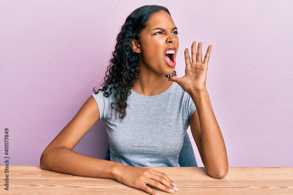 Young african american girl wearing casual clothes sitting on the table shouting and screaming loud to side with hand on mouth. communication concept. Young african american girl wearing casual clothes sitting on the table shouting and screaming loud to side with hand on mouth. communication concept.