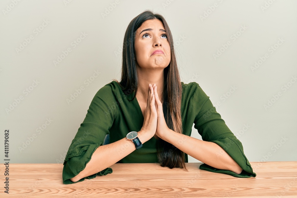 Young brunette woman wearing casual clothes sitting on the table begging and praying with hands together with hope expression on face very emotional and worried. begging. Young brunette woman wearing casual clothes sitting on the table begging and praying with hands together with hope expression on face very emotional and worried. begging.