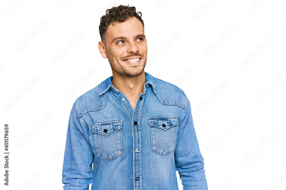 Young hispanic man wearing casual denim jacket looking away to side with smile on face, natural expression. laughing confident. Young hispanic man wearing casual denim jacket looking away to side with smile on face, natural expression. laughing confident.
