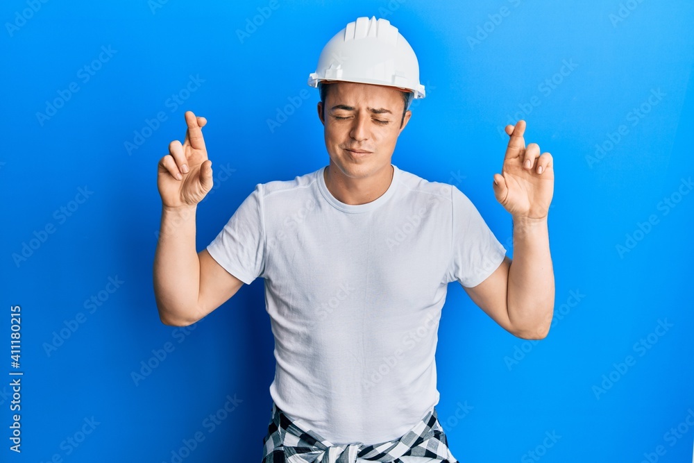 Handsome young man wearing builder uniform and hardhat gesturing finger crossed smiling with hope and eyes closed. luck and superstitious concept. Handsome young man wearing builder uniform and hardhat gesturing finger crossed smiling with hope and eyes closed. luck and superstitious concept.