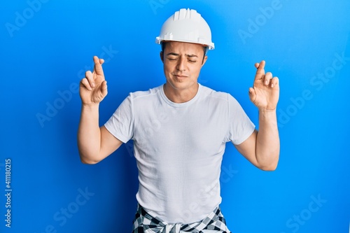 Handsome young man wearing builder uniform and hardhat gesturing finger crossed smiling with hope and eyes closed. luck and superstitious concept.