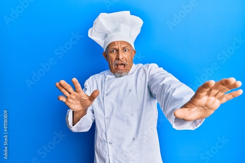 Middle age grey-haired man wearing professional cook uniform and hat afraid and terrified with fear expression stop gesture with hands, shouting in shock. panic concept.